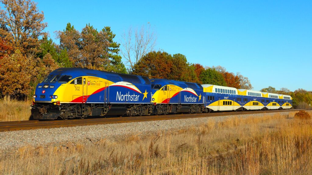 Blue and yellow train travels along autumn colored trees.