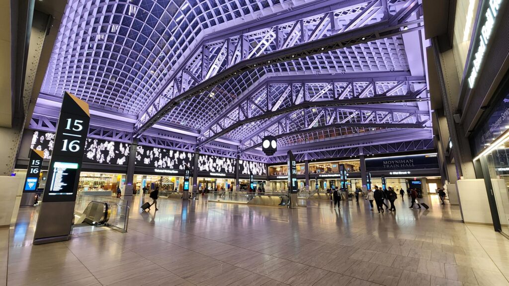 The dramatic vaulted ceiling of Moynihan Train Hall showcasing intricate steel arch trusses and geometric coffered panels illuminated in purple and lavender light. Travelers move through the spacious concourse below while departure boards and the Moynihan Train Hall sign mark the far wall.