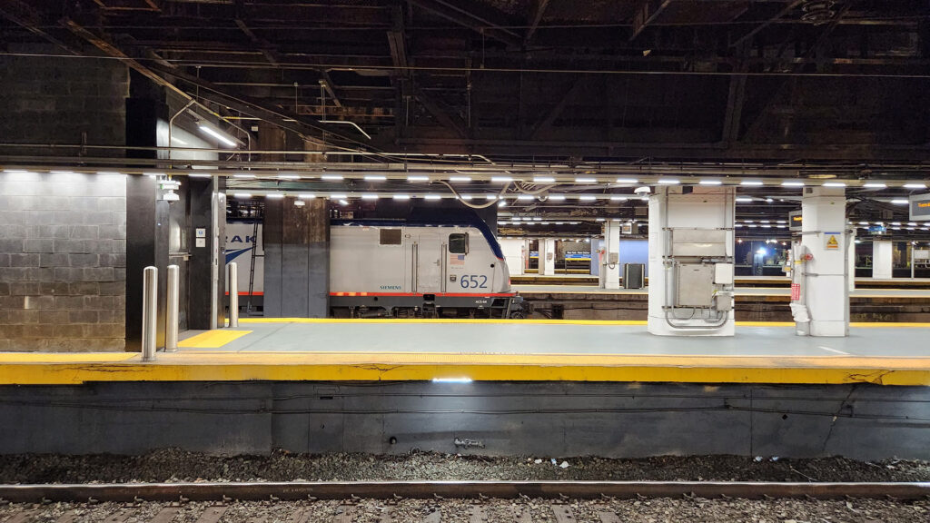 Platform view at New York Penn Station showing Amtrak ACS-64 electric locomotive #652 at rest beside the bright yellow safety stripe. The industrial platform infrastructure reveals exposed ceiling beams, concrete columns, and utilitarian lighting that defines the working heart of Northeast Corridor rail service.