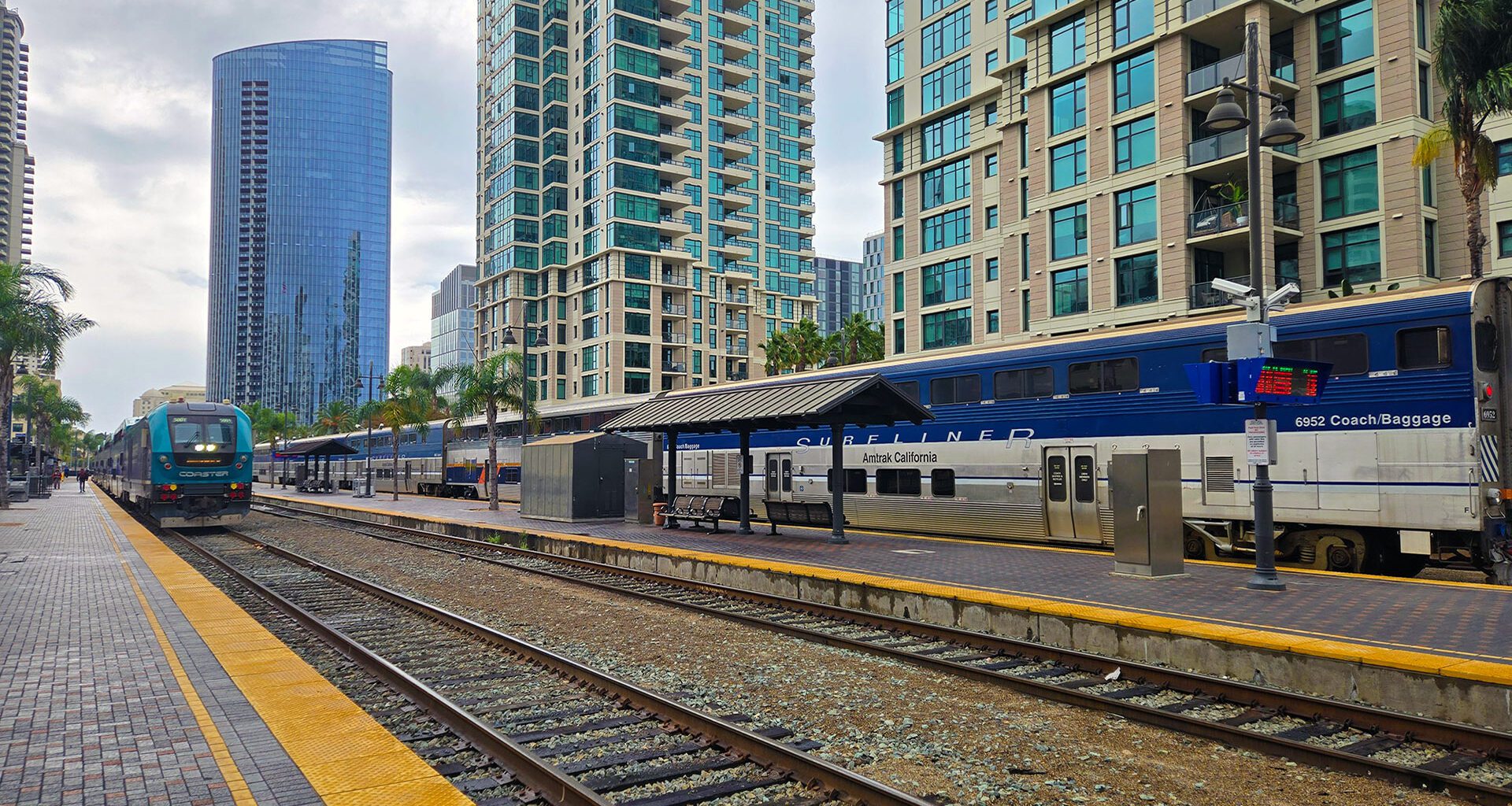 Two trains waiting at the platform with taller glass buildings in the background.