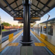 View from the center of a LA Metro light rail station platform showing two silver Metro trains on opposite sides. Yellow tactile warning strips line the platform edges. Digital arrival information boards hang from the covered platform structure. Warm golden hour sunlight illuminates the scene, with trees and residential buildings visible in the background.