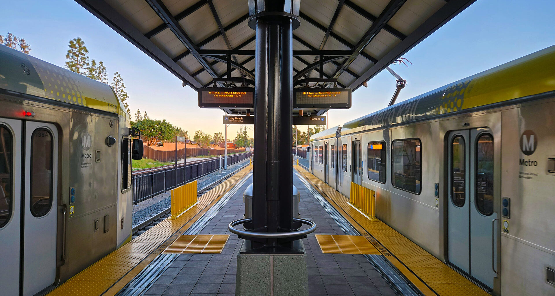 View from the center of a LA Metro light rail station platform showing two silver Metro trains on opposite sides. Yellow tactile warning strips line the platform edges. Digital arrival information boards hang from the covered platform structure. Warm golden hour sunlight illuminates the scene, with trees and residential buildings visible in the background.