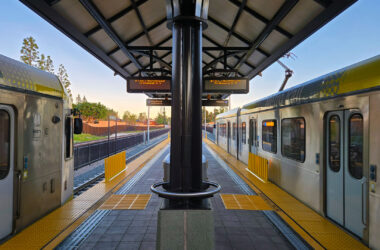 View from the center of a LA Metro light rail station platform showing two silver Metro trains on opposite sides. Yellow tactile warning strips line the platform edges. Digital arrival information boards hang from the covered platform structure. Warm golden hour sunlight illuminates the scene, with trees and residential buildings visible in the background.
