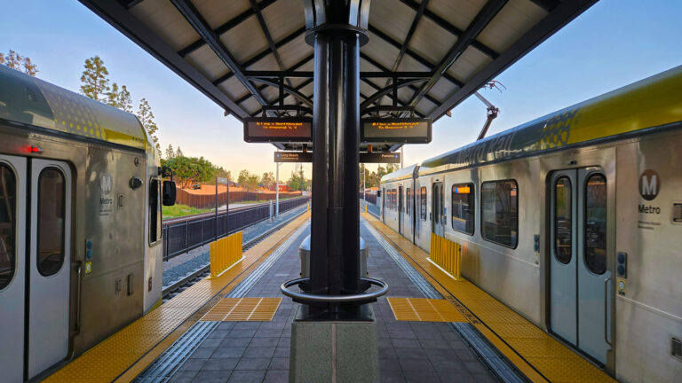 View from the center of a LA Metro light rail station platform showing two silver Metro trains on opposite sides. Yellow tactile warning strips line the platform edges. Digital arrival information boards hang from the covered platform structure. Warm golden hour sunlight illuminates the scene, with trees and residential buildings visible in the background.