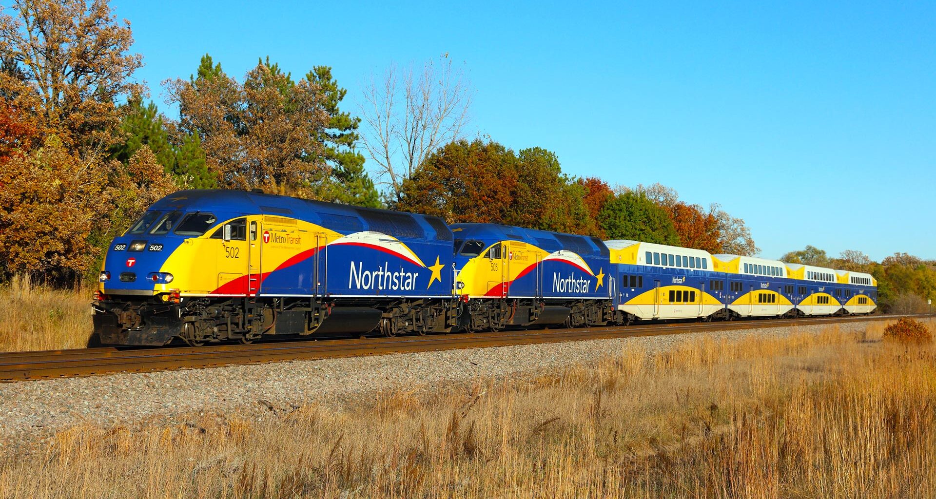 Blue and yellow train travels along autumn colored trees.