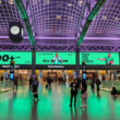 Moynihan Train Hall's main concourse bathed in dramatic green uplighting, with travelers crossing the polished floor beneath soaring vaulted ceilings. Numbered ticketed waiting rooms line the walls, large advertising displays glow above the entrances, and an art deco-style clock hangs suspended from the coffered ceiling supported by exposed steel arches.
