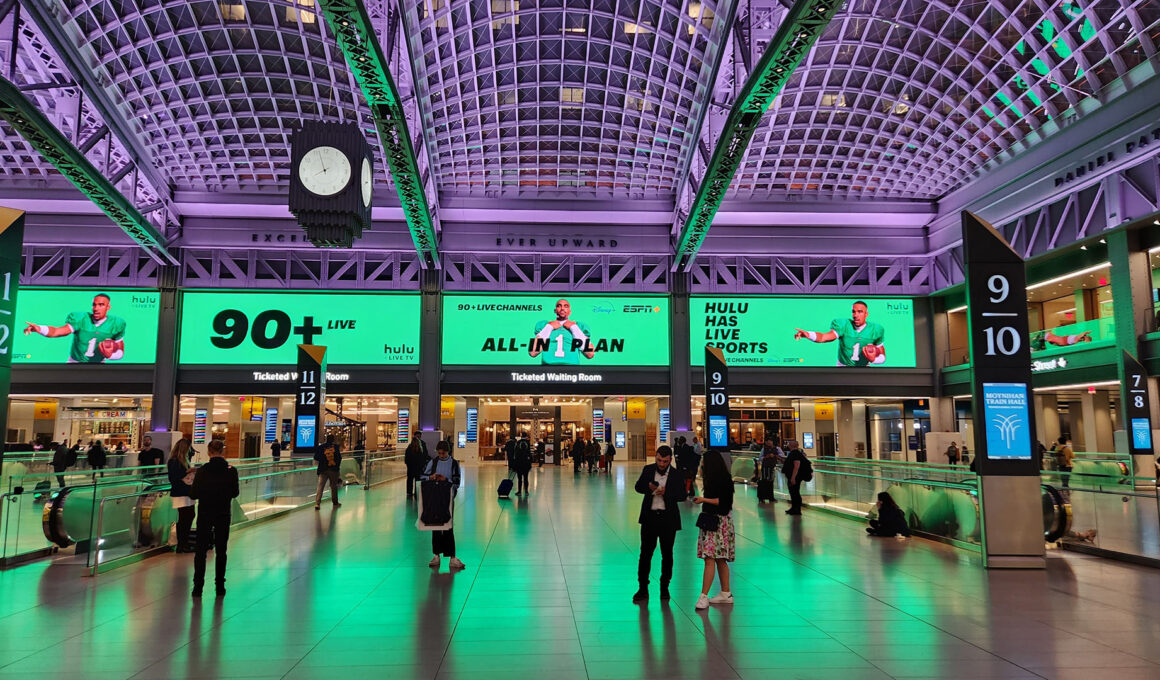 Moynihan Train Hall's main concourse bathed in dramatic green uplighting, with travelers crossing the polished floor beneath soaring vaulted ceilings. Numbered ticketed waiting rooms line the walls, large advertising displays glow above the entrances, and an art deco-style clock hangs suspended from the coffered ceiling supported by exposed steel arches.