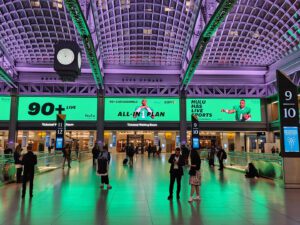 Moynihan Train Hall's main concourse bathed in dramatic green uplighting, with travelers crossing the polished floor beneath soaring vaulted ceilings. Numbered ticketed waiting rooms line the walls, large advertising displays glow above the entrances, and an art deco-style clock hangs suspended from the coffered ceiling supported by exposed steel arches.
