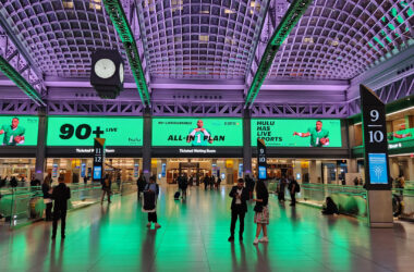 Moynihan Train Hall's main concourse bathed in dramatic green uplighting, with travelers crossing the polished floor beneath soaring vaulted ceilings. Numbered ticketed waiting rooms line the walls, large advertising displays glow above the entrances, and an art deco-style clock hangs suspended from the coffered ceiling supported by exposed steel arches.