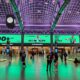 Moynihan Train Hall's main concourse bathed in dramatic green uplighting, with travelers crossing the polished floor beneath soaring vaulted ceilings. Numbered ticketed waiting rooms line the walls, large advertising displays glow above the entrances, and an art deco-style clock hangs suspended from the coffered ceiling supported by exposed steel arches.