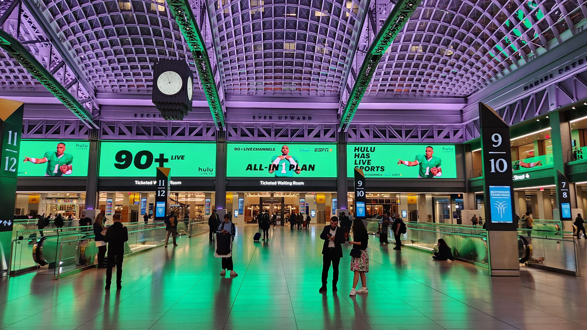 Moynihan Train Hall's main concourse bathed in dramatic green uplighting, with travelers crossing the polished floor beneath soaring vaulted ceilings. Numbered ticketed waiting rooms line the walls, large advertising displays glow above the entrances, and an art deco-style clock hangs suspended from the coffered ceiling supported by exposed steel arches.