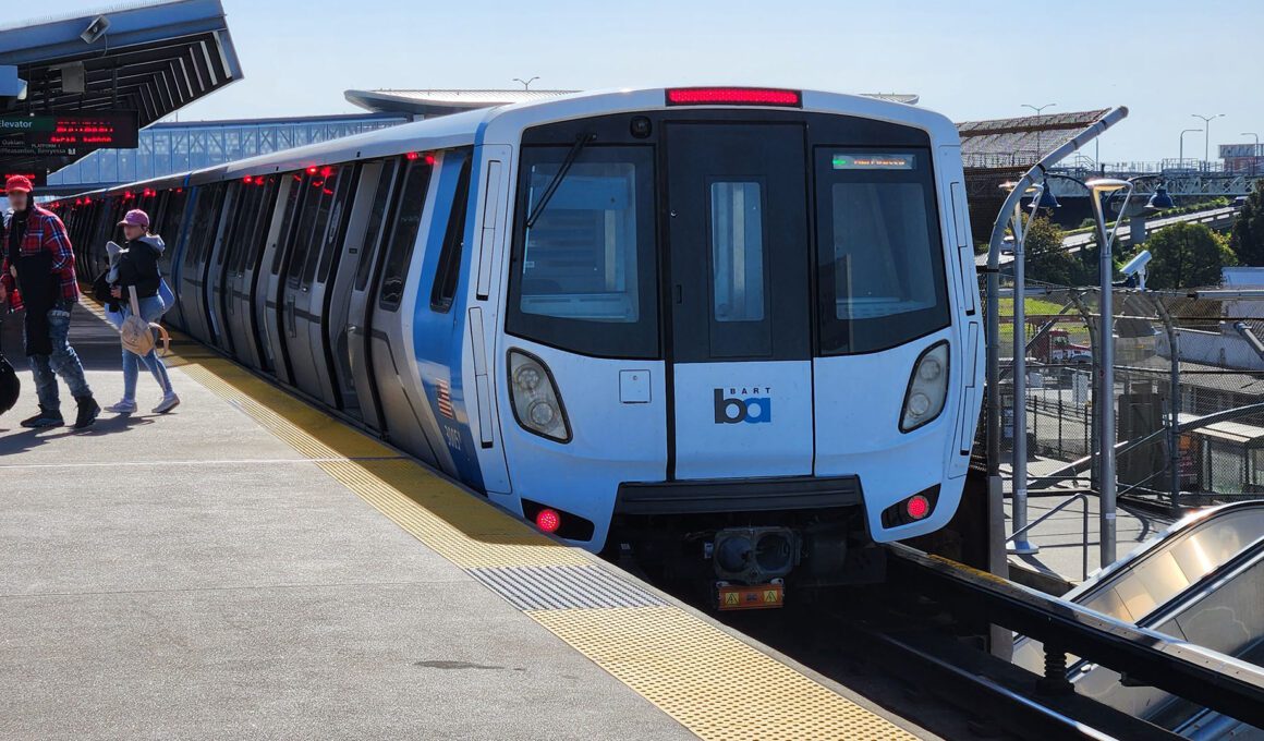 A BART train sits at an outdoor elevated station platform on a clear day, with passengers walking alongside the blue and white Fleet of the Future car.