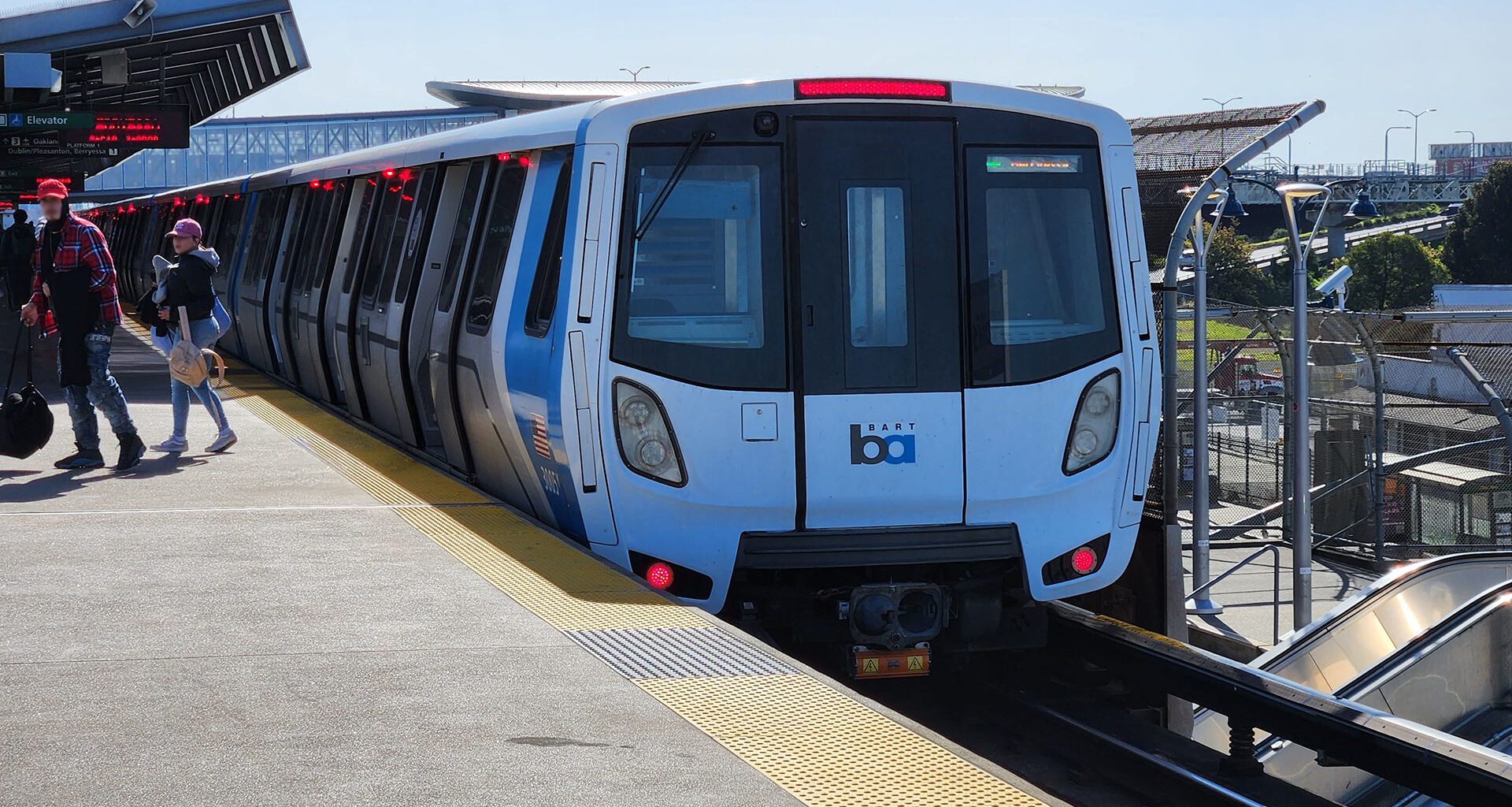 A BART train sits at an outdoor elevated station platform on a clear day, with passengers walking alongside the blue and white Fleet of the Future car.