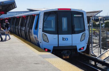 A BART train sits at an outdoor elevated station platform on a clear day, with passengers walking alongside the blue and white Fleet of the Future car.