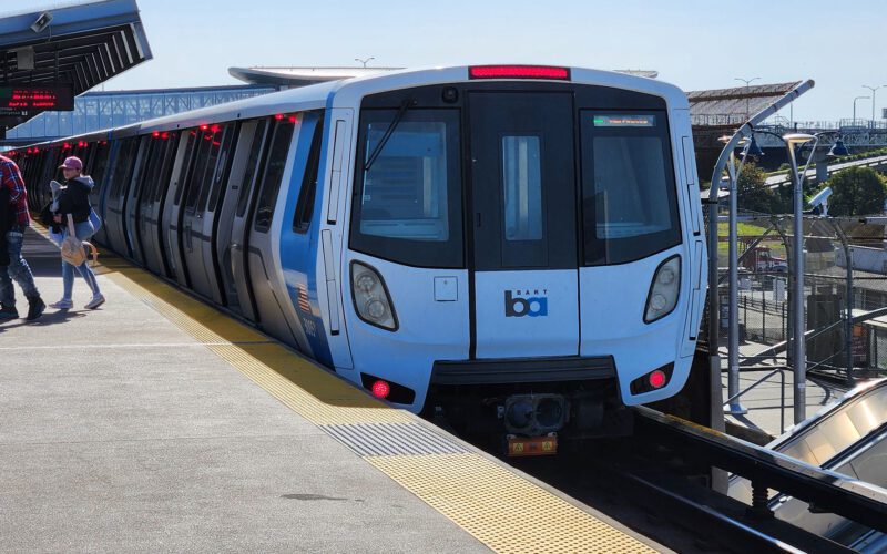 A BART train sits at an outdoor elevated station platform on a clear day, with passengers walking alongside the blue and white Fleet of the Future car.