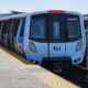 A BART train sits at an outdoor elevated station platform on a clear day, with passengers walking alongside the blue and white Fleet of the Future car.