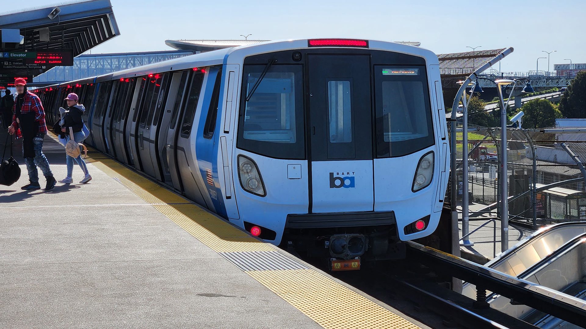 A BART train sits at an outdoor elevated station platform on a clear day, with passengers walking alongside the blue and white Fleet of the Future car.