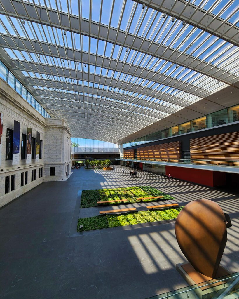 - Vector+Vista Bright glass-roofed atrium with greenery beds, wooden benches, and a large abstract sculpture at the foreground