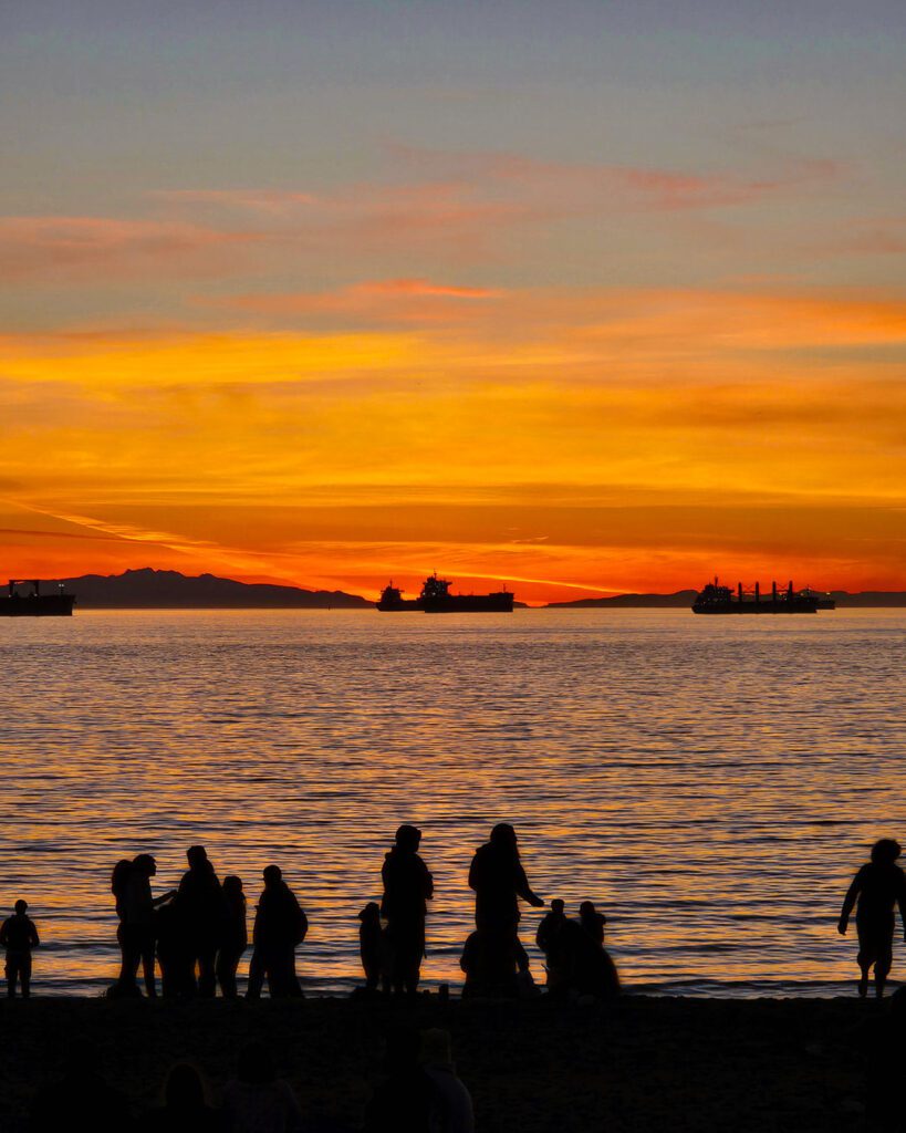 - Vector+Vista Silhouetted people on a beach at sunset with a orange sky and ships on the horizon.