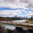 View from an Amtrak train departing Philadelphia, with the skyline and highway bridge visible along the Schuylkill River, representing the rail corridor a future Reading to Philadelphia line would extend.