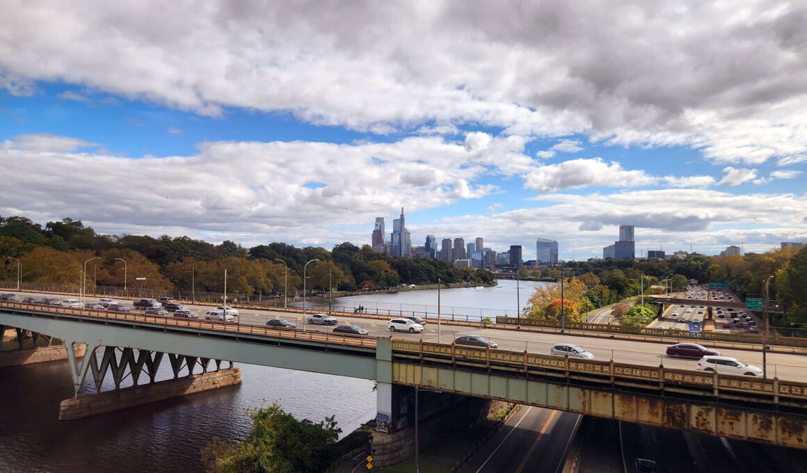 View from an Amtrak train departing Philadelphia, with the skyline and highway bridge visible along the Schuylkill River, representing the rail corridor a future Reading to Philadelphia line would extend.