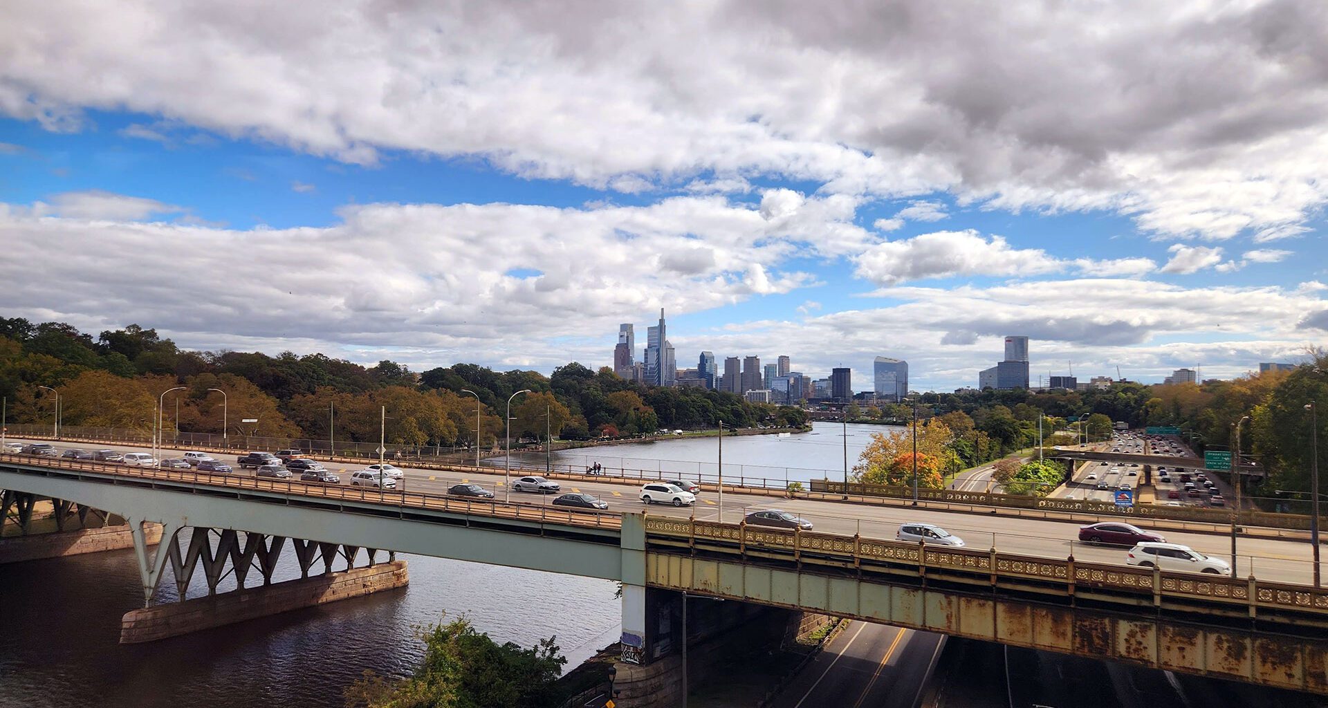 View from an Amtrak train departing Philadelphia, with the skyline and highway bridge visible along the Schuylkill River, representing the rail corridor a future Reading to Philadelphia line would extend.