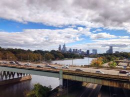 View from an Amtrak train departing Philadelphia, with the skyline and highway bridge visible along the Schuylkill River, representing the rail corridor a future Reading to Philadelphia line would extend.