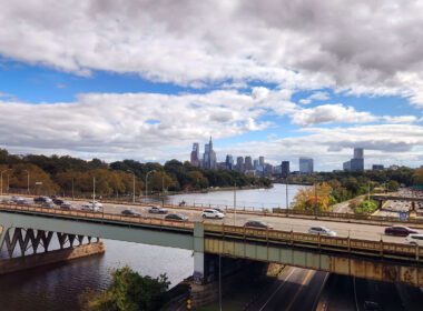 View from an Amtrak train departing Philadelphia, with the skyline and highway bridge visible along the Schuylkill River, representing the rail corridor a future Reading to Philadelphia line would extend.