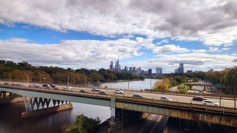 View from an Amtrak train departing Philadelphia, with the skyline and highway bridge visible along the Schuylkill River, representing the rail corridor a future Reading to Philadelphia line would extend.