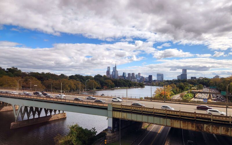 View from an Amtrak train departing Philadelphia, with the skyline and highway bridge visible along the Schuylkill River, representing the rail corridor a future Reading to Philadelphia line would extend.